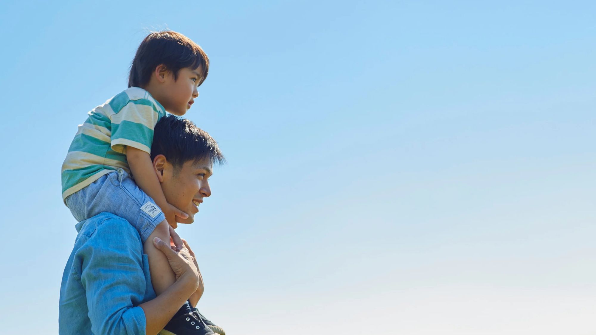 A father carrying his son on his shoulders outdoors under a blue sky